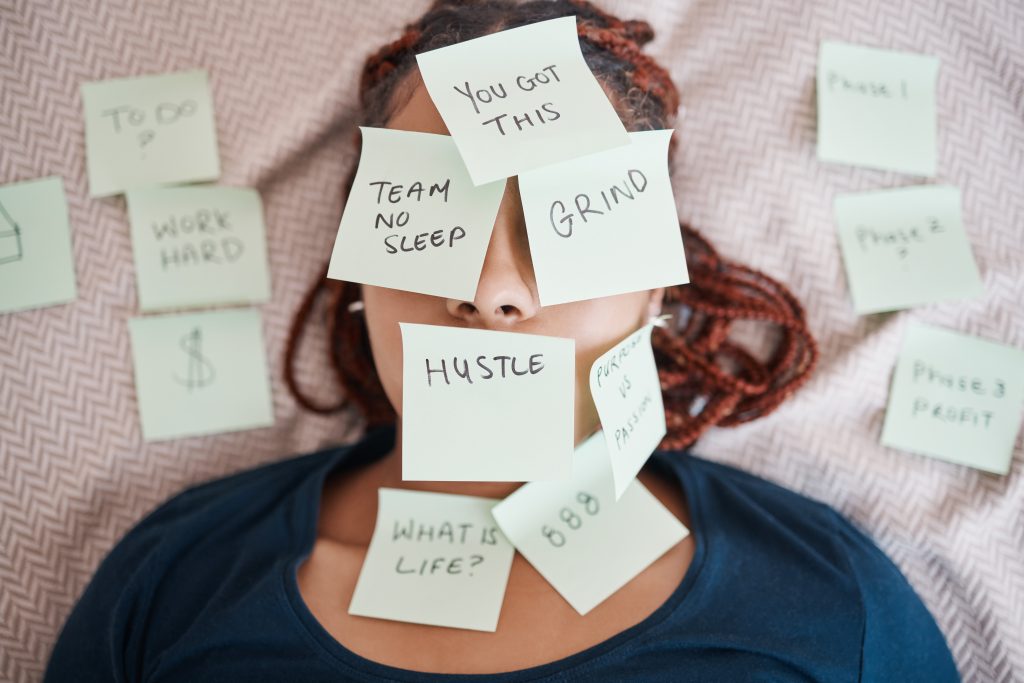 Person lying down with multiple sticky notes on their face, each displaying motivational or work-related phrases like "Hustle," "Grind," and "No Sleep"—a snapshot of procrastination and the hidden cost of always pushing harder.